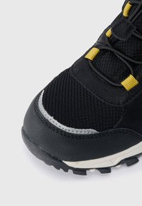 Μαύρο και κίτρινο παπούτσι πεζοπορίας με άκρο από διχτυωτό υλικό και καουτσούκ, διαθέτοντας παχύ πέλμα με αυλακώσεις και κίτρινες θηλιές κορδονιών σε λευκό φόντο.