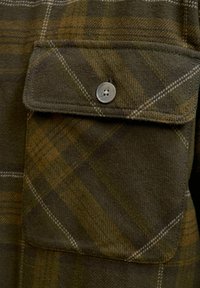 Grünes kariertes Flanellhemd mit einer Brusttasche und einem silbernen Knopf, weicher Textur und kontrastierenden weißen Nähten an den Kanten.