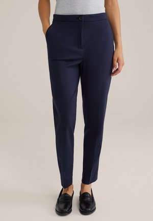 Woman wearing navy slim-fit trousers with front button and black loafers, standing against light background.