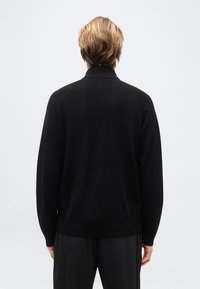 Personne blonde debout de dos, portant un pull noir à manches longues avec des détails en panneaux côtelés et un pantalon noir, sur un fond clair.