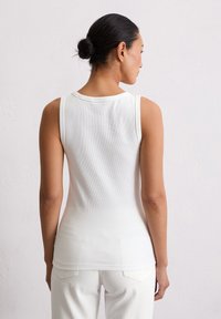 Weißes ärmelloses, geripptes Tanktop mit enger Passform und rundem Ausschnitt, kombiniert mit hellen Hosen vor einem neutralen Hintergrund.