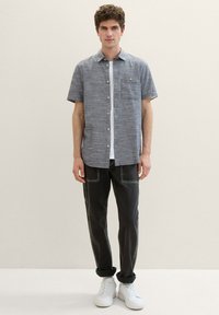 Cămașă din in albastră cu mânecă scurtă și buzunar pe piept, asortată cu pantaloni din denim închis și adidași albi. Outfitul are un croi lejer.