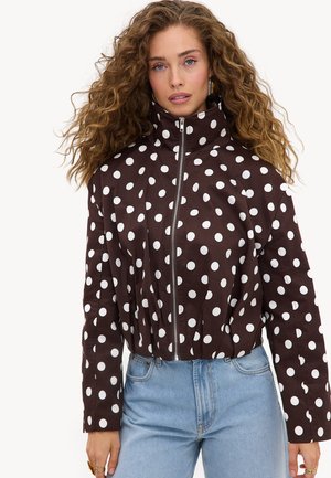 Femme aux cheveux bouclés portant une veste marron à col haut avec des pois blancs et un jean bleu clair, face à l’avant.