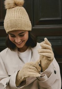 Beige strikket hat med en blød kvast, parret med matchende ribstrikkede handsker. Teksturen er blød med lodrette striber over det hele.