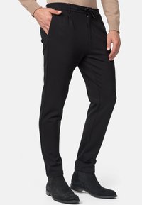 Mężczyzna ubrany w dopasowane czarne spodnie z elastycznym paskiem i czarne zamszowe buty Chelsea, ręce spoczywające blisko kieszeni.