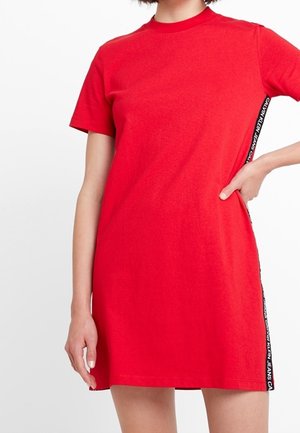 Frau trägt ein knielanges, kurzärmeliges, rotes Kleid mit schwarzen Seitenpartien, die weißen Calvin Klein Jeans Logo-Text zeigen.