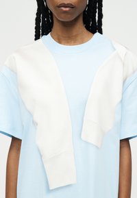 Άτομο που φοράει ανοιχτό μπλε κοντομάνικο πουκάμισο με λευκή μπλούζα φούτερ περασμένη στους ώμους, εμφανιζόμενο από τους ώμους μέχρι το πηγούνι.