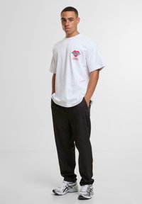 White cotton t-shirt with short sleeves, featuring blue and red graphic text. Paired with black pants and silver athletic shoes.