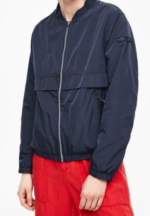 Person wearing a navy blue zip-up bomber jacket with elastic cuffs and red pants, standing against a plain white background.