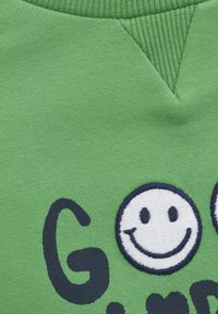 Grünes Baumwollhemd mit einem marineblauen gestickten Smiley-Gesicht und schwarzem Text. Rippkragen mit einem dreieckigen Designdetail.