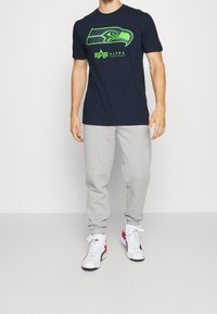 Námořnická modrá tričko s krátkým rukávem se zeleným logem Seahawks, doplněné o šedé tepl áky a bílé tenisky s černými a červenými akcenty.