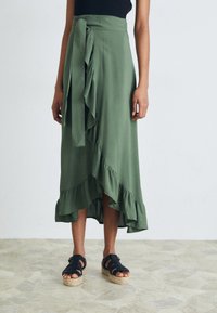 Gonna avvolgente di colore verde oliva con fondo a volant, un dettaglio con cravatta in vita, abbinata a sandali espadrillas neri con cinturini su un pavimento Texturato.