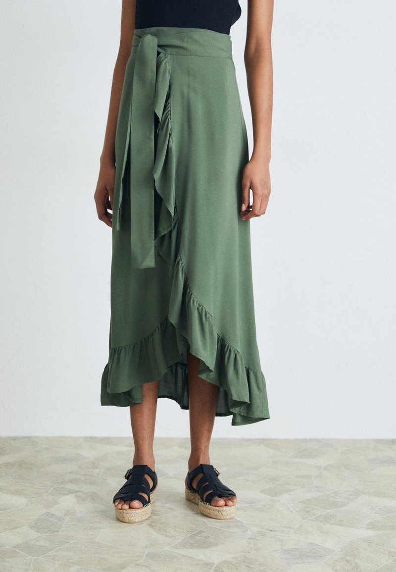 Gonna avvolgente di colore verde oliva con fondo a volant, un dettaglio con cravatta in vita, abbinata a sandali espadrillas neri con cinturini su un pavimento Texturato.