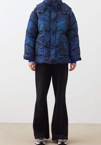 Giacca puffer blu navy con un motivo floreale in tonalità di blu e verde, dotata di un alto colletto e tasche frontali, abbinata a pantaloni neri.