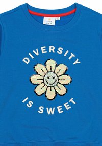 Plavi dukserica s crvenim izrezom, koja ima veliki, šljokičasti cvjetni uzorak. Natpis glasi "DIVERZITET JE SLATKO" u bijeloj boji.