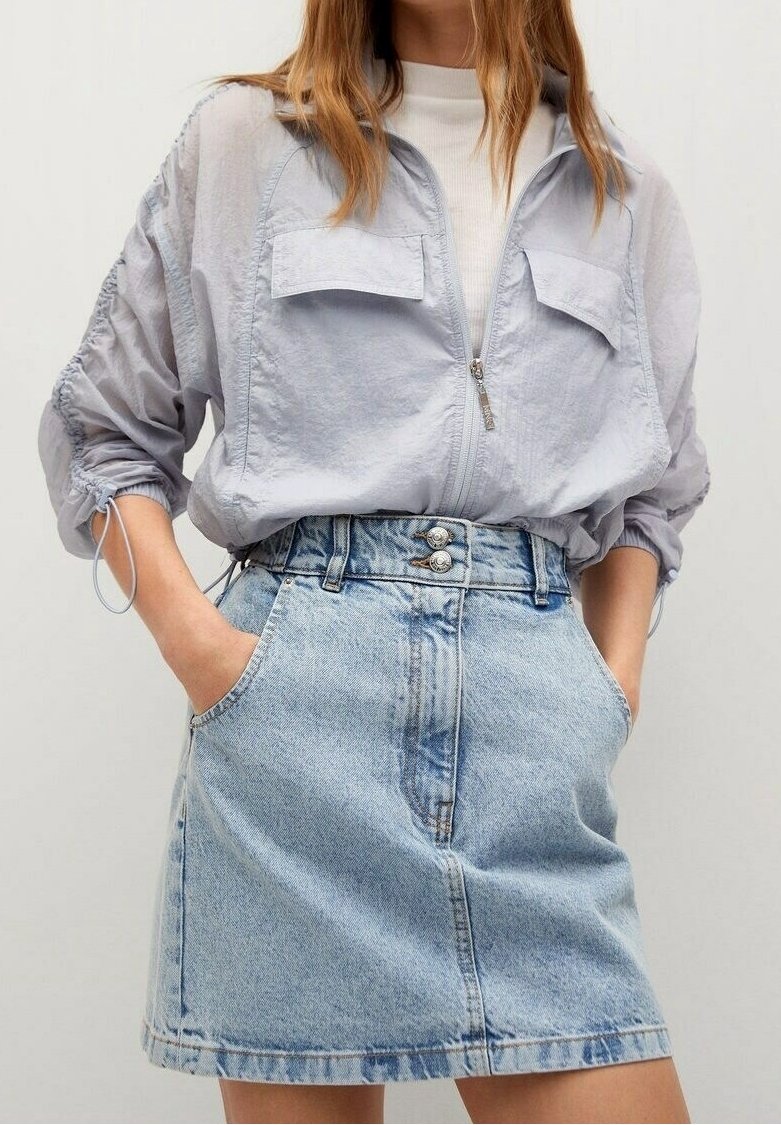 Femme portant une veste zippée gris clair sur un haut blanc et une jupe en jean bleu clair taille haute, les mains dans les poches.