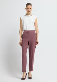 Witte mouwloze top met een bewerkte halslijn, gecombineerd met smalle bordeauxrode broeken. Het model draagt bruine hoge hakken. De achtergrond is eenvoudig.