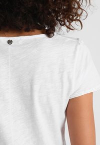 Camiseta blanca de manga corta con cuello redondo, que presenta un pequeño logo metálico en la parte de atrás y una tela sutilmente texturizada.