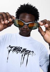Slnečné okuliare s textúrovanými zeleno-oranžovými šošovkami; čierny rám. Nosené osobou v bielom tričku s čiernym graffiti štýlom textu.