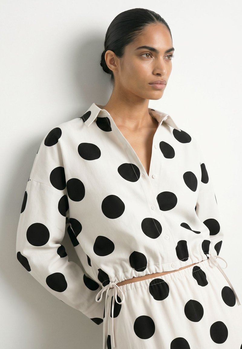 Femme aux cheveux foncés en chignon portant une chemise blanche à manches longues et un pantalon à gros pois noirs, debout contre un mur blanc.