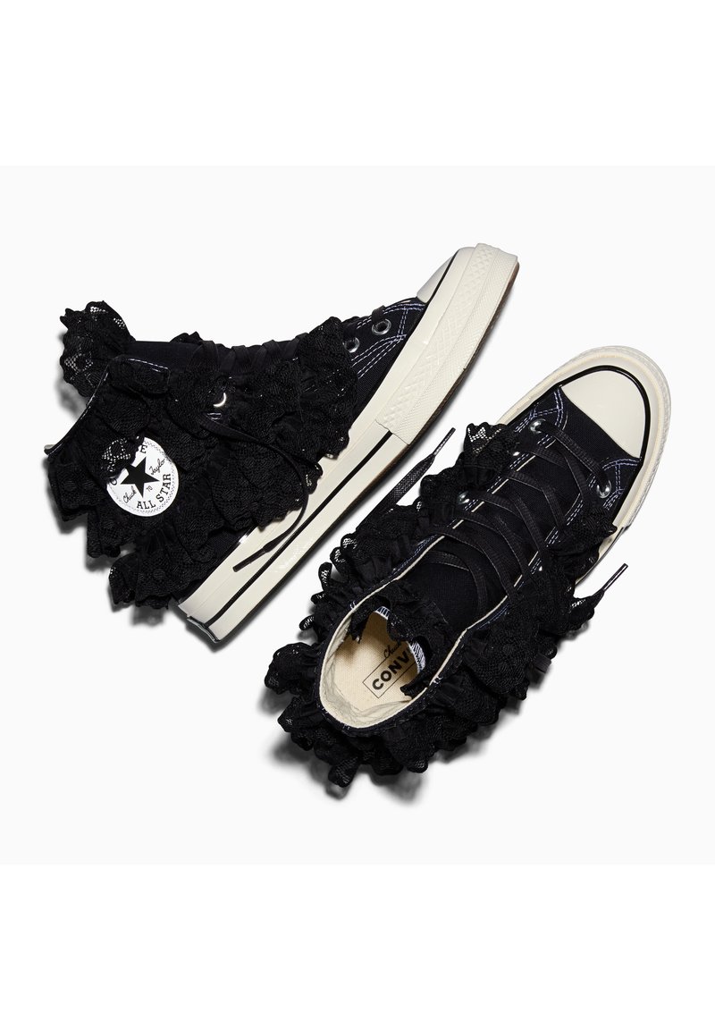 Zapatillas Converse negras de caña alta con suelas y punteras blancas, decoradas con volantes de encaje negro alrededor de los tobillos y la lengüeta.