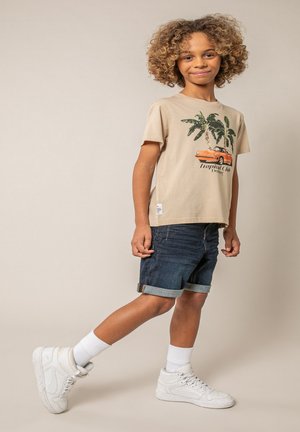 Enfant souriant aux cheveux bouclés portant un t-shirt beige à imprimé tropical, un short en denim foncé retroussé, des chaussettes blanches et des baskets blanches, posant sur beige.