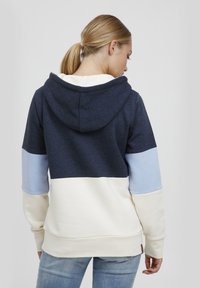 Hettegenser i marineblå, lyseblå og kremfarge. Har en fargeblokkeringsdesign med myk tekstur og ribbede mansjetter.
