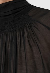Blusa nera trasparente con colletto alto, dettagli arricciati sullo scollo e tessuto morbido e fluido con una sottile texture.