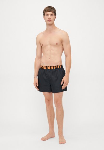 Ein oberkörperfreier junger Mann steht barfuß in schwarzen Badeshorts mit orangefarbenem Logo-Bund vor einem schlichten weißen Hintergrund.