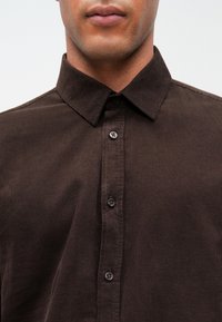 Ciemnobrązowa koszula zapinana na guziki, wykonana z fakturowanej tkaniny, z wykładanym kołnierzykiem i widocznymi guzikami z przodu.