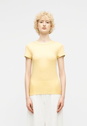 Νεαρό άτομο με κοντά σγουρά ξανθά μαλλιά, που φοράει ανοιχτό κίτρινο κοντομάνικο πουκάμισο και χαλαρά λευκά παντελόνια, στέκεται μπροστά σε απλό φόντο.