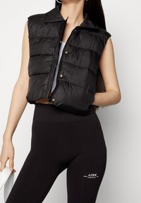 Schwarze gepolsterte Weste mit kragenförmigem Ausschnitt, goldenen Druckknöpfen und ärmellosem Design, kombiniert mit schwarzen gerippten Leggings mit einem Logo.