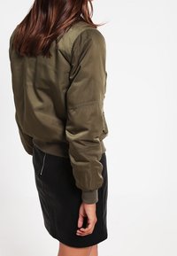 Olivově zelená bomber bunda s hladkým, lesklým povrchem, žebrovanými manžetami a páskem, a bočními zipovými kapsami. Nositá přes černou sukni.