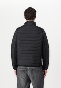 Black insulated jacket featuring horizontal quilted panels, stand collar, and contrasting fabric on sides. Worn with gray jeans.