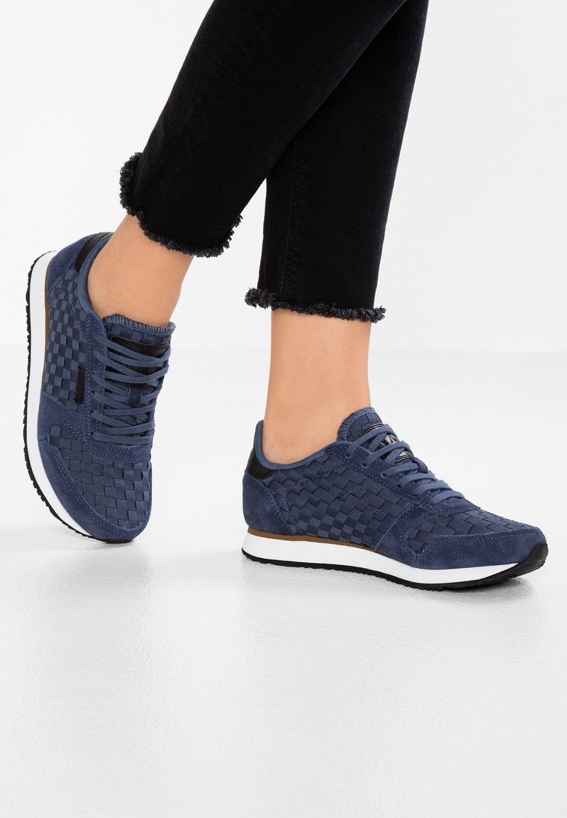Personne portant un pantalon court noir et des baskets tissées bleu marine avec des semelles blanches, debout sur un sol blanc brillant.
