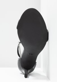 Chaussure noire à talon haut avec un bout pointu, tige en cuir texturé et semelle lisse avec des stries ; accent en chaîne argentée sur la bride.