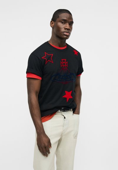 Νέος άνδρας που φοράει μαύρο t-shirt με κόκκινα αστέρια και έμβλημα, από πάνω από κόκκινο πουκάμισο, σε συνδυασμό με ανοιχτόχρωμο παντελόνι μπροστά σε απλό φόντο.