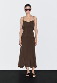 Hnědé midi šaty se spagetti ramínky, volánovými vrstvami, měkkou tkaninou a rozšířeným spodním lemem. V kombinaci s černými plochými sandály a nadrozměrnými slunečními brýlemi.
