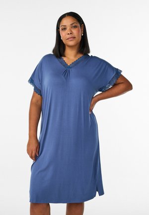 Žena stojaca s rukou na bedre, oblečená v modrých šatách po kolená s čipkovaným lemom na rukávoch a výstrihu, na bielom pozadí.