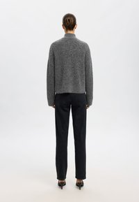 Grå texturerad stickad tröja med hög krage, tillsammans med raka svarta jeans och svarta klackar, sedd bakifrån.