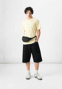 T-shirt gialla con testo "NARANJAS", marsupio nero, pantaloni corti di denim scuro, calze grigie e sneakers multicolori, su sfondo bianco.