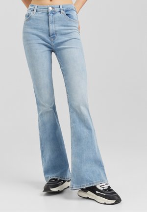 Person trägt hellblaue Schlagjeans und schwarz-weiße Turnschuhe, steht vor einem schlichten grauen Hintergrund.