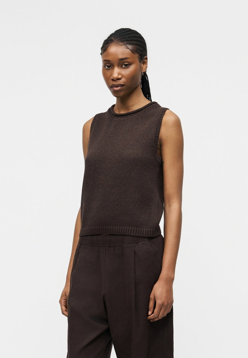 Γυναίκα με κοτσίδες που φοράει αμάνικο σκούρο καφέ πλεκτό τοπ και φαρδιά σκούρα καφέ παντελόνια μπροστά από απλό ανοιχτό φόντο.