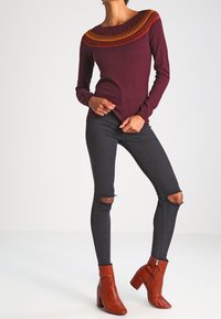 Pull bordeaux avec un col orné de motifs complexes orange et jaune, associé à un jean skinny noir déchiré et des bottines marron à talons.