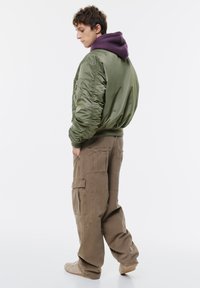 Zöld bomber dzseki, alatta lila kapucnis pulóver, oversized barna cargo nadrággal, amely több zsebbel rendelkezik.