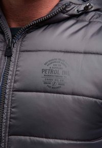 Närbild av en person som bär en grå vadderad jacka med dragkedja och tryckt text "PETROL IND" på bröstområdet.