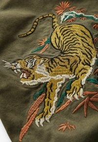Broderad patch med en detaljerad tiger i gult och svart på olivgrön tyg, omgiven av färgglad lövverk och blommig dekoration.