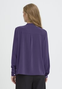 Dolga rukava vijolična bluzica z ohlapnim krojem, gumbe na manšetah, gladko teksturo in detajlom na hrbtni strani. Enostaven dizajn brez vzorcev.