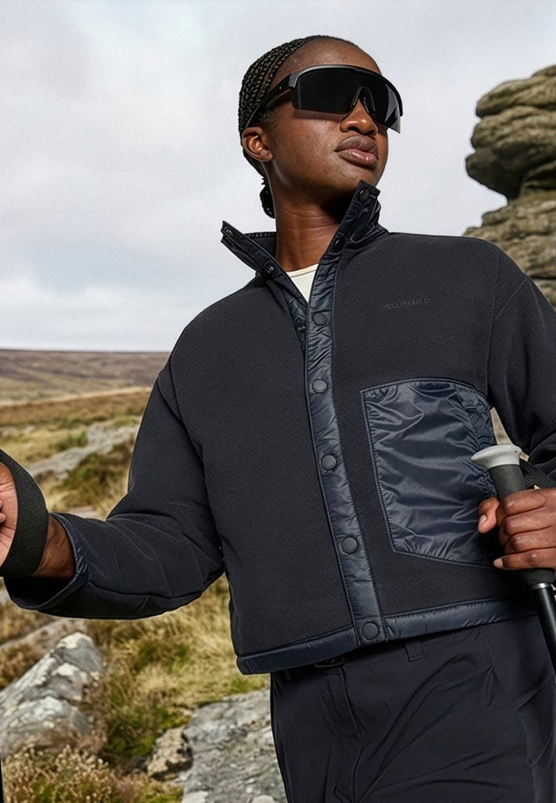 Personne portant une veste d’extérieur sombre et de grandes lunettes de soleil, tenant des bâtons de marche, debout sur un terrain rocheux avec un ciel nuageux en arrière-plan.