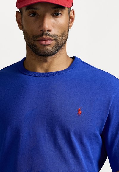 Βασιλικό μπλε μπλουζάκι με μακριά μανίκια, ριγωτό λαιμό και μικρό κόκκινο κεντητό λογότυπο στο στήθος. Το ύφασμα φαίνεται λείο και ελαφρύ.